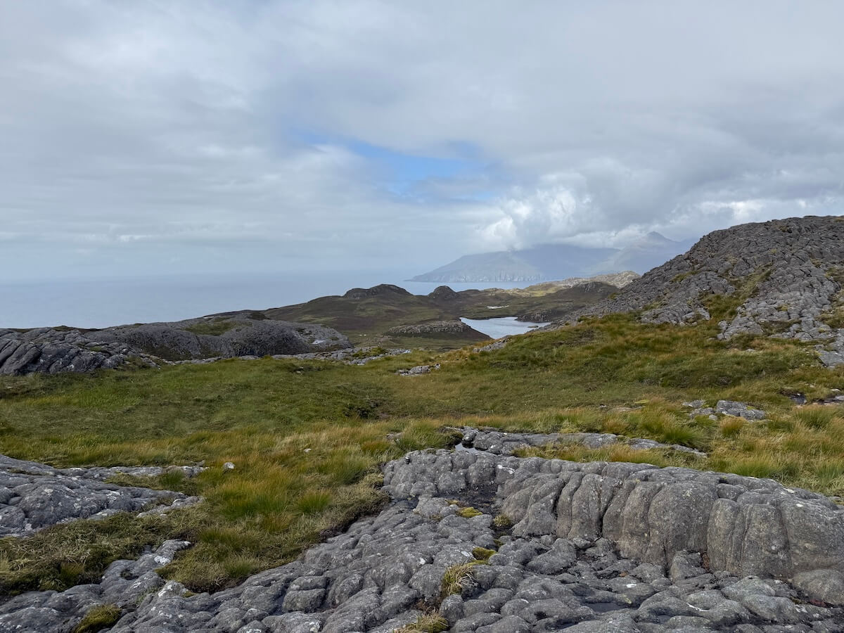 Rocky foreground leads across grassy hills towards a distant coastline and low mountains partly hidden by cloud. The wide landscape feels remote under a moody sky.