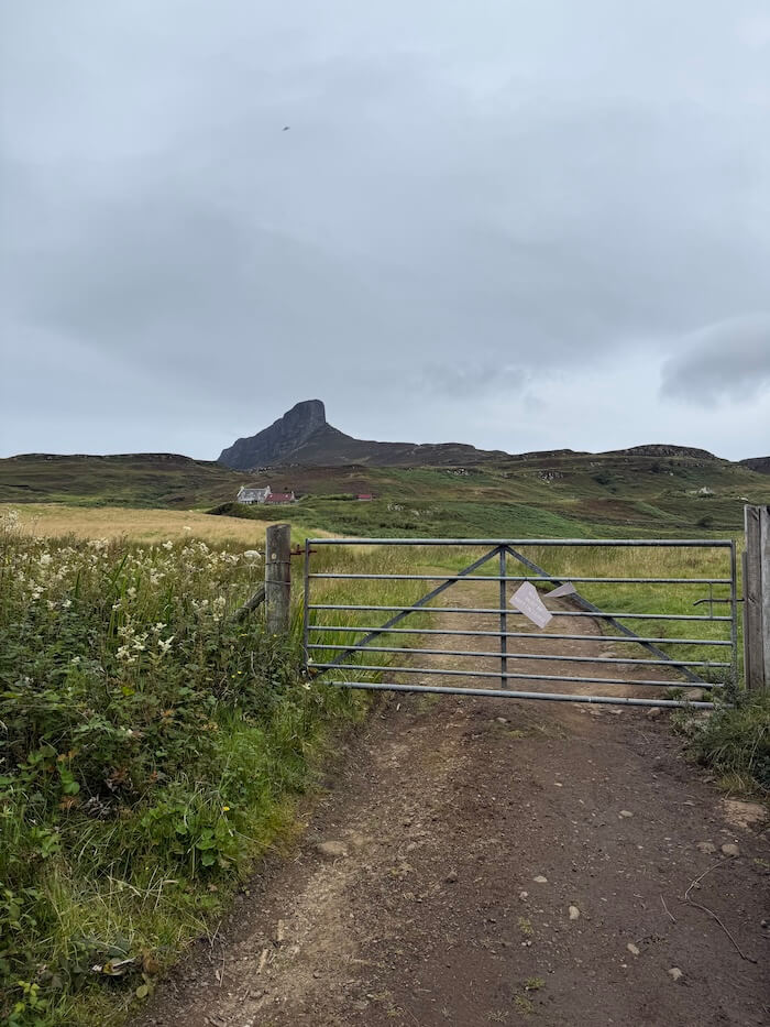 Metal farm gate blocks a dirt track leading across open fields towards a distinctive rocky hill in the distance. The landscape stretches out under a grey sky.