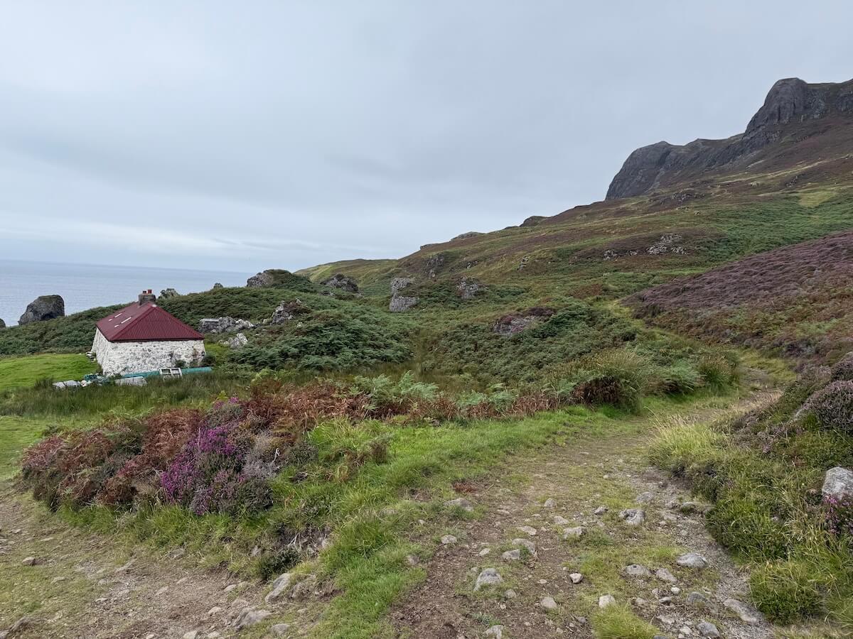 Winding grassy path leads past a small stone cottage with a red roof, set among heather and shrubs on a coastal hillside. The sea and rocky slopes rise in the background under a grey sky.