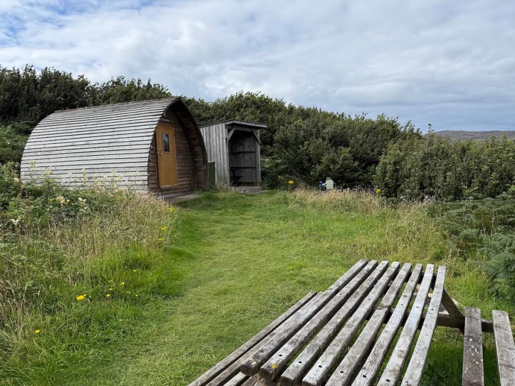Small wooden camping pod and open shelter set beside a grassy path surrounded by tall wild plants and dense shrubs. A weathered picnic table in the foreground adds to the quiet rural campsite setting.