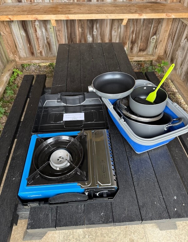 Portable camp stove, frying pan, nested pots, and a bright green spatula arranged on a black picnic table under a wooden shelter. The setup shows compact outdoor cooking gear ready for a camping meal.