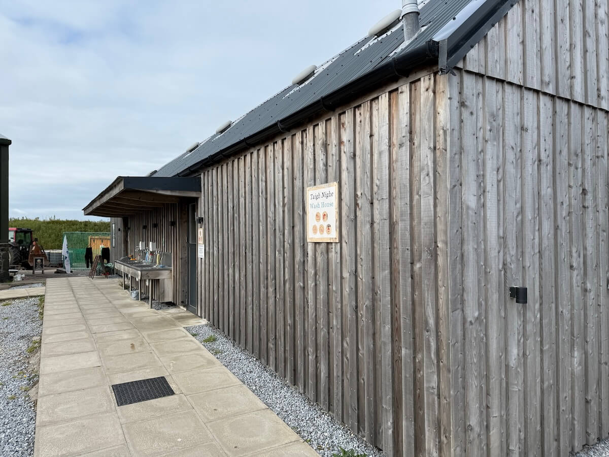 Long wooden wash house building beside a paved campsite path with outdoor sinks under a covered section. A sign on the wall reads "Taigh Nighe Wash House".