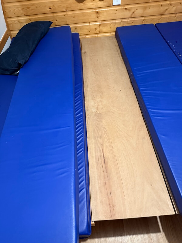 Two narrow blue sleeping mats separated by a removable wooden center panel inside the camping pod. The arrangement shows how the sleeping area can be divided.