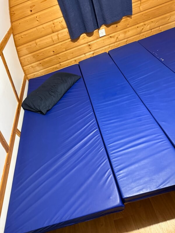 Blue sleeping mats spread across the pod floor to create a wider bed setup. A single dark pillow rests on one side beneath a curtained window.