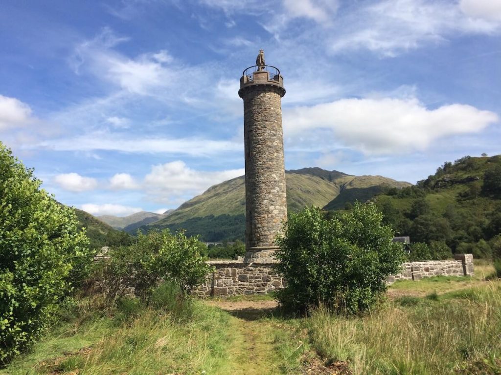 Glenfinnan Monument rising above a grassy valley in the Scottish Highlands with rugged green hills, stone walls and scattered trees beneath a bright blue sky with drifting clouds.