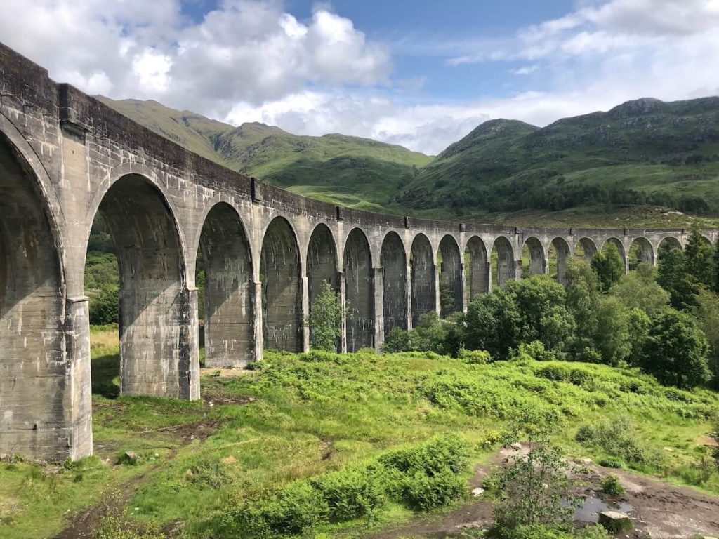 Glenfinnan Viaduct curving across a green Highland valley with stone arches, rolling hills and bright summer clouds overhead.