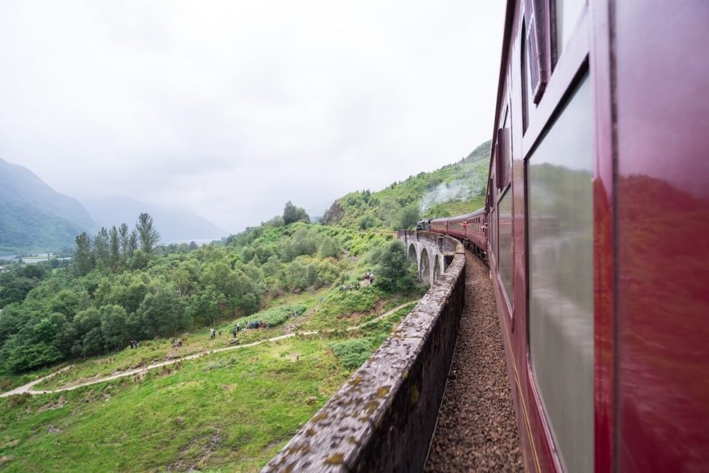 View from the Jacobite steam train as it curves across the Glenfinnan Viaduct through the Scottish Highlands with green hills, drifting steam and walkers taking photographs below the bridge on a cloudy day.