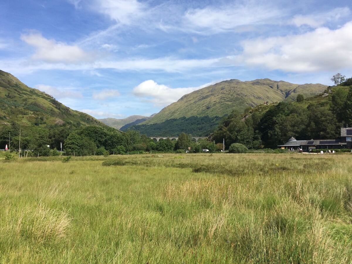 Wide grassy field near Glenfinnan with mountains and the Glenfinnan Viaduct visible in the distance under a bright blue sky.