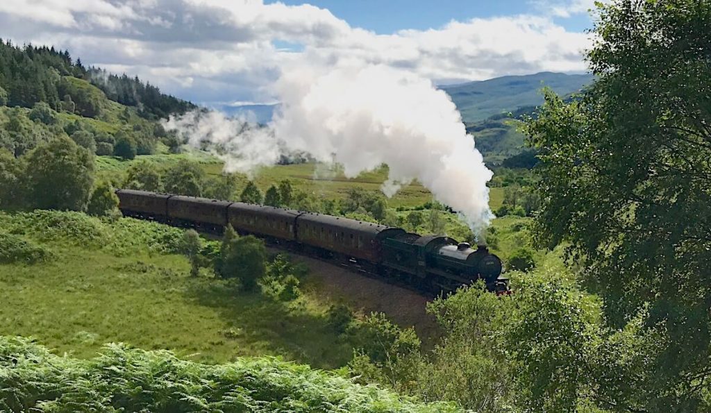 Steam train travelling through a lush green valley with rolling hills and scattered trees in the background. Thick white steam rises above the carriages beneath a bright sky filled with large clouds.