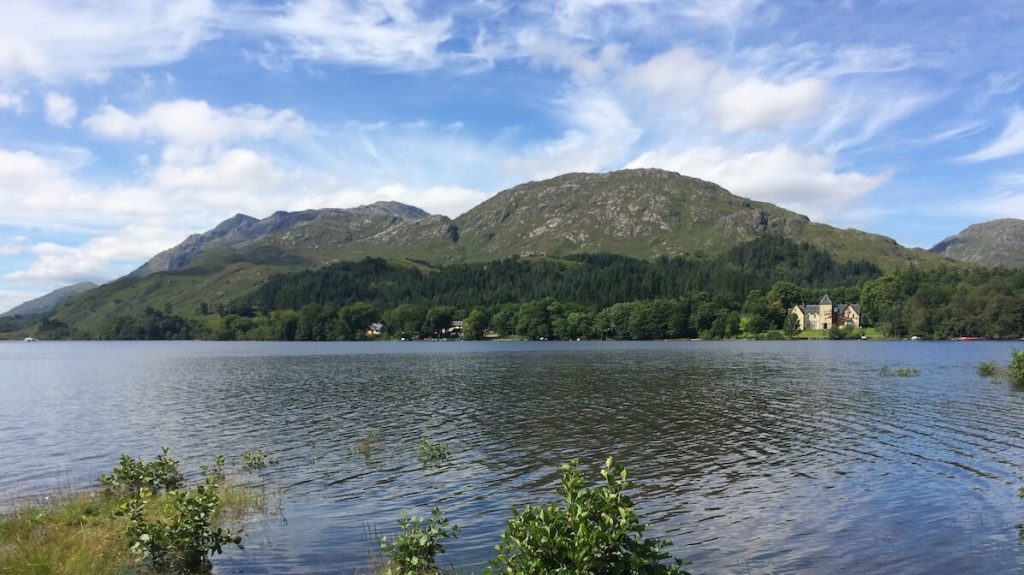View across Loch Shiel towards green mountains and a lochside house beneath a partly cloudy sky.