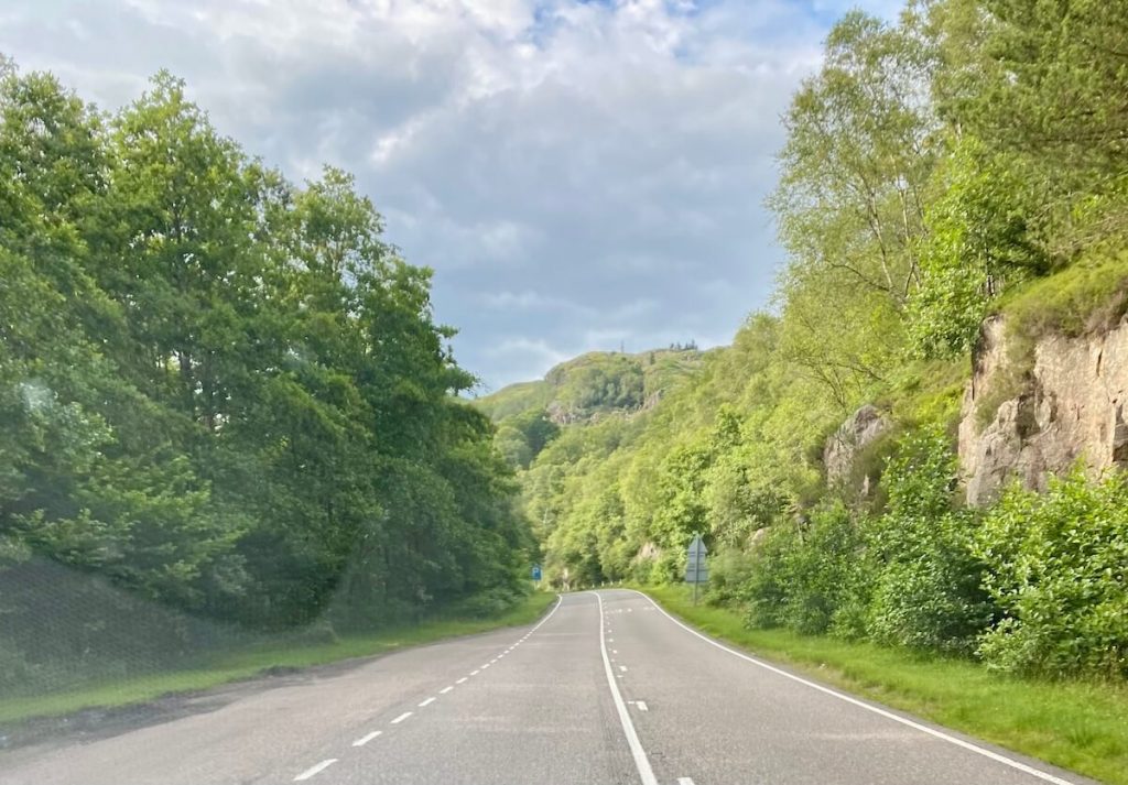 Winding Highland road lined with green trees and rocky hillsides on a cloudy summer day in Scotland.