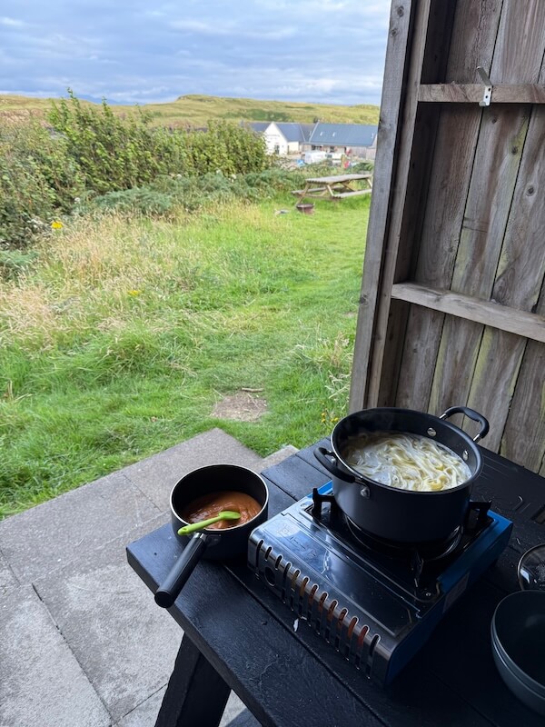 Pot of pasta cooking on a portable stove beside a small saucepan of red sauce on a sheltered outdoor table. The open view looks onto grass hedges and distant campsite buildings beyond the cooking area.