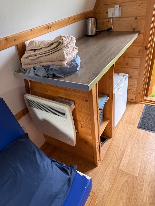 Compact wooden camping pod interior with folded towels and bedding on a countertop above a small shelf and mini fridge. A wall heater and the edge of a blue mattress show the simple sleeping and storage setup.