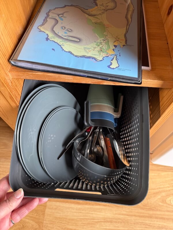 Open storage basket inside the pod filled with plates mugs cutlery and cooking utensils. A folded map rests on the wooden shelf above the basket.