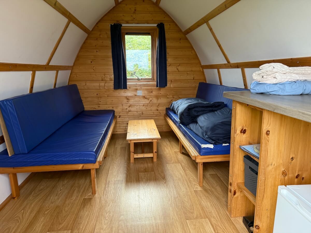 Full interior view of the wooden camping pod with bench seating on one side a made bed on the other and a small table between them. A window at the back brings light into the compact cabin space.
