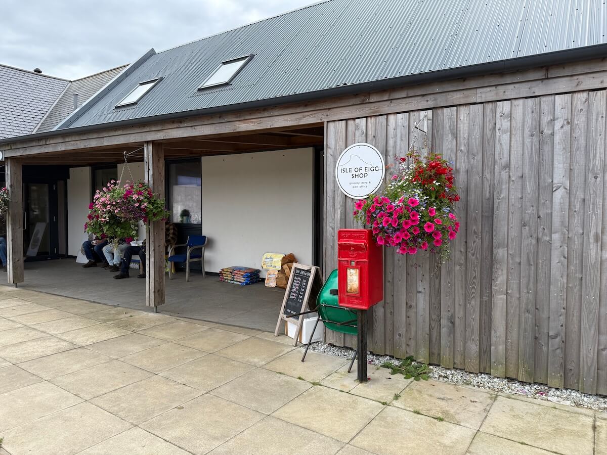 Wooden storefront for the Isle of Eigg Shop with hanging flower baskets a bright red post box and a covered entrance with chairs outside. A round sign reads "ISLE OF EIGG SHOP grocery store and post office".