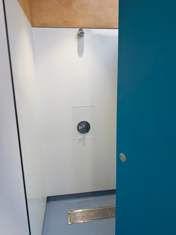 Private shower stall with white walls a blue door a wall mounted shower head and a floor drain. The space appears simple and practical for campsite facilities.