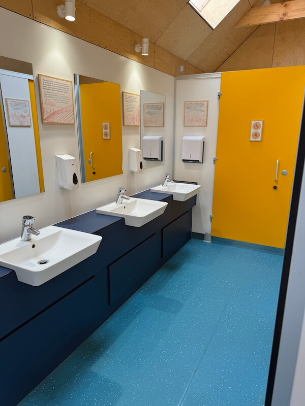 Bright shared washroom with three sinks mirrors soap dispensers hand dryers and yellow stall doors. Accessibility signage appears on one stall door and framed information notices hang above the sinks.
