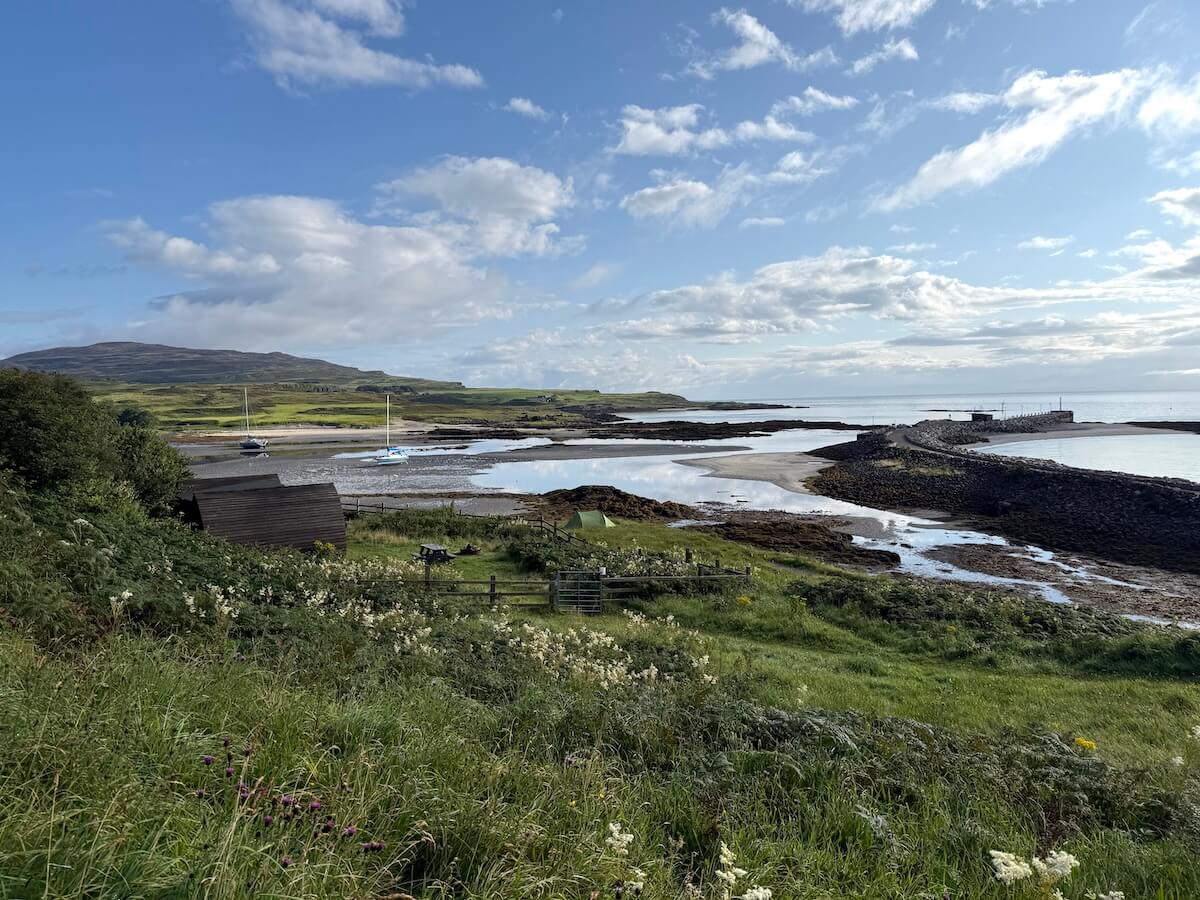 Coastal camping pod view across grassy wildflower slopes toward a sheltered bay at low tide with two small sailboats resting near the shore. Rocky seawalls and distant green hills sit beneath a bright blue sky with scattered clouds.