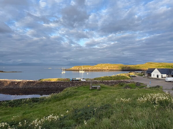 Calm coastal harbour viewed from a grassy rise with a stone sea wall boats on the water and a pale building beside the shore. Evening light brightens the low island across the bay beneath layered clouds.
