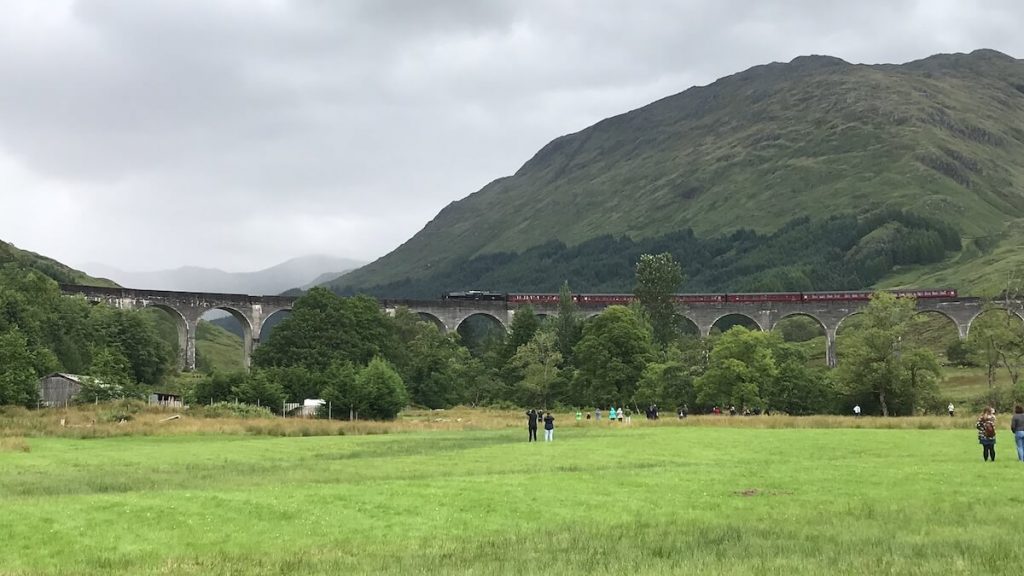 Jacobite steam train crossing the Glenfinnan Viaduct with mountains and grassy fields in the background.